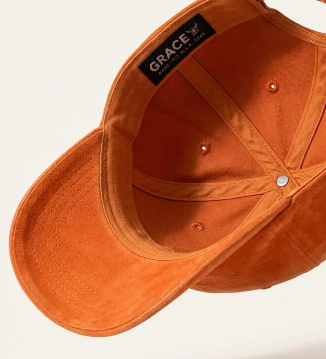 An orange suede baseball cap shown from an angled top-down view, floating against a light beige background with a soft shadow underneath. The brand name 'GRACE' is visible on a black label stitched inside the crown.