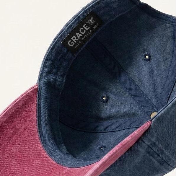 A dual-tone baseball cap shown floating at an angle against a light cream background. The crown and brim are made of a textured navy blue fabric, while the underside of the brim is a contrasting dusty rose pink.