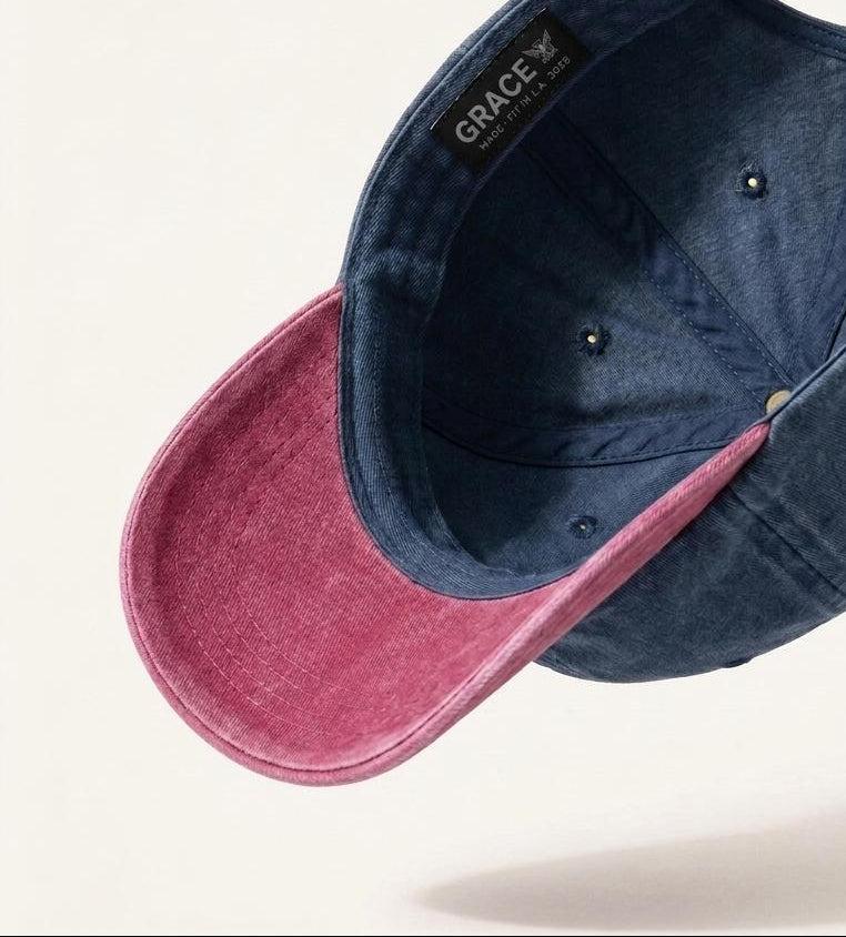 A dual-tone baseball cap shown floating at an angle against a light cream background. The crown and brim are made of a textured navy blue fabric, while the underside of the brim is a contrasting dusty rose pink.