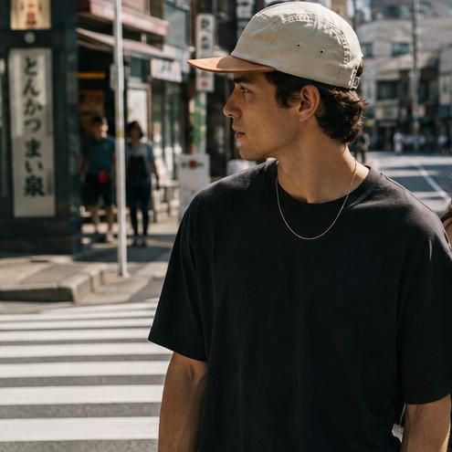 A man and a woman walking side-by-side on a sunny city street. The man is wearing a black t-shirt and a beige baseball cap, while the woman wears a black sleeveless top and sunglasses.