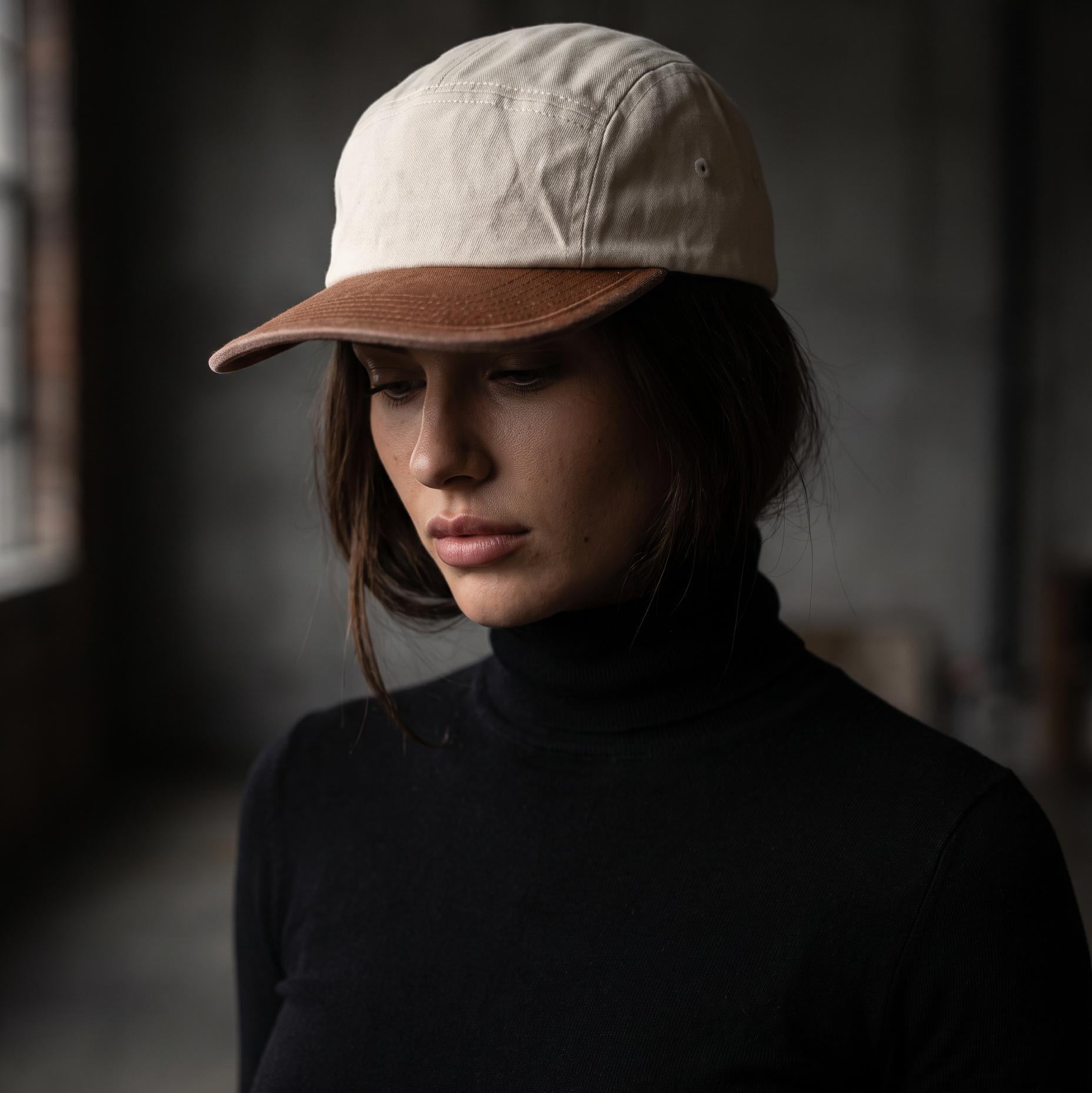 A woman wearing a light beige cotton baseball cap with a brown suede visor, paired with a black turtleneck sweater, set against a dark, moody indoor background.
