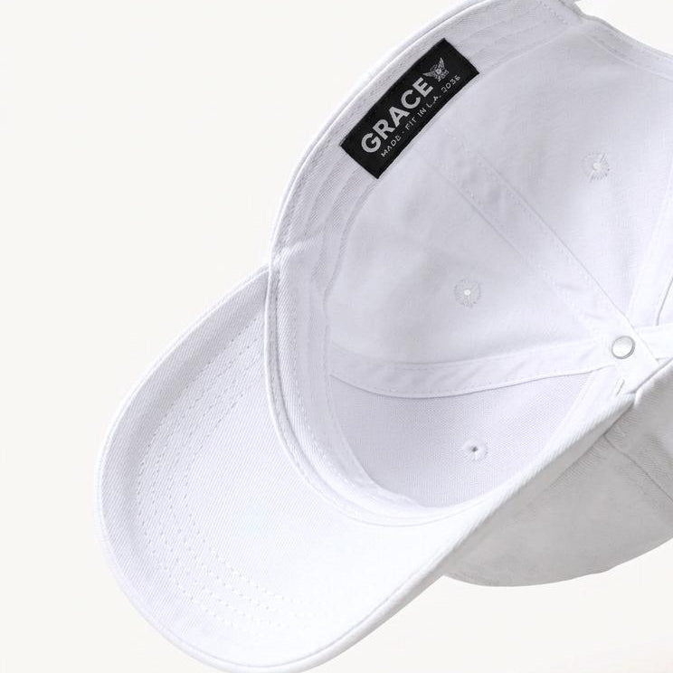 A white baseball cap shown floating at an angle against a light cream background, with a black interior label reading GRACE and a small gold metal accent near the back strap.