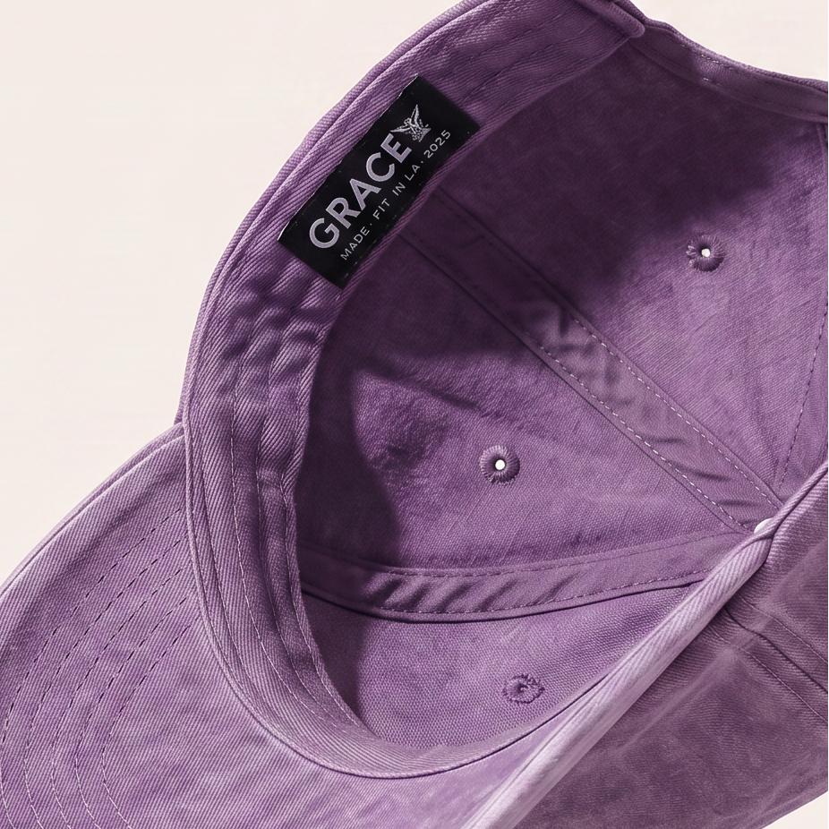A purple washed cotton baseball cap floating at an angle against a warm off-white background, showing the interior with a black 'GRACE' brand label.