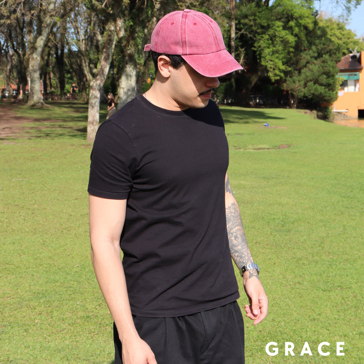 Man wearing a black t-shirt and pink cap in a park setting with 'GRACE' branding.