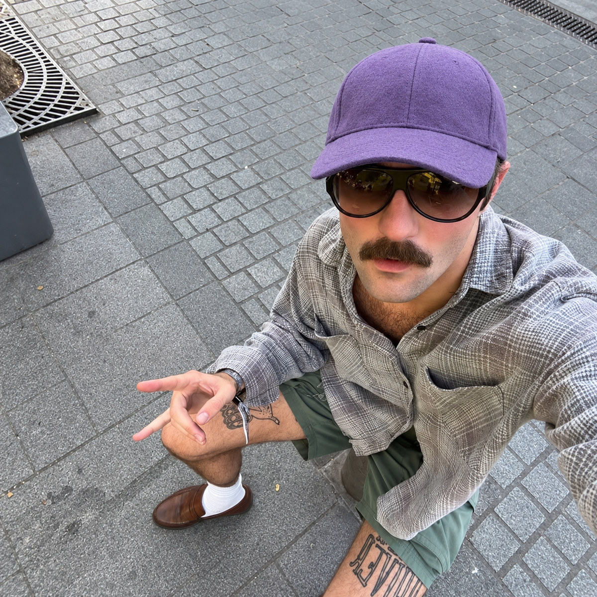 A man wearing a purple baseball cap, black sunglasses, a gray plaid button-up shirt, olive green shorts, and brown loafers, taking a selfie on a gray paved outdoor walkway.