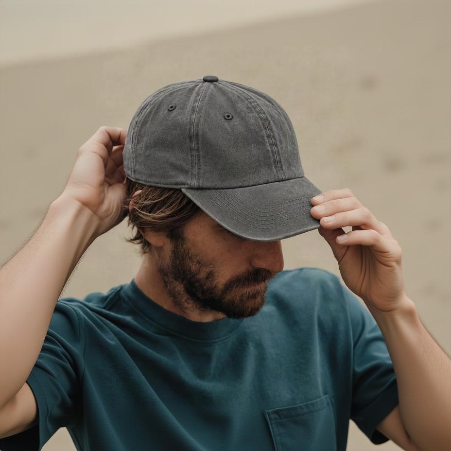A man wearing a washed charcoal gray baseball cap and a teal t-shirt, adjusting the brim of the cap with both hands.