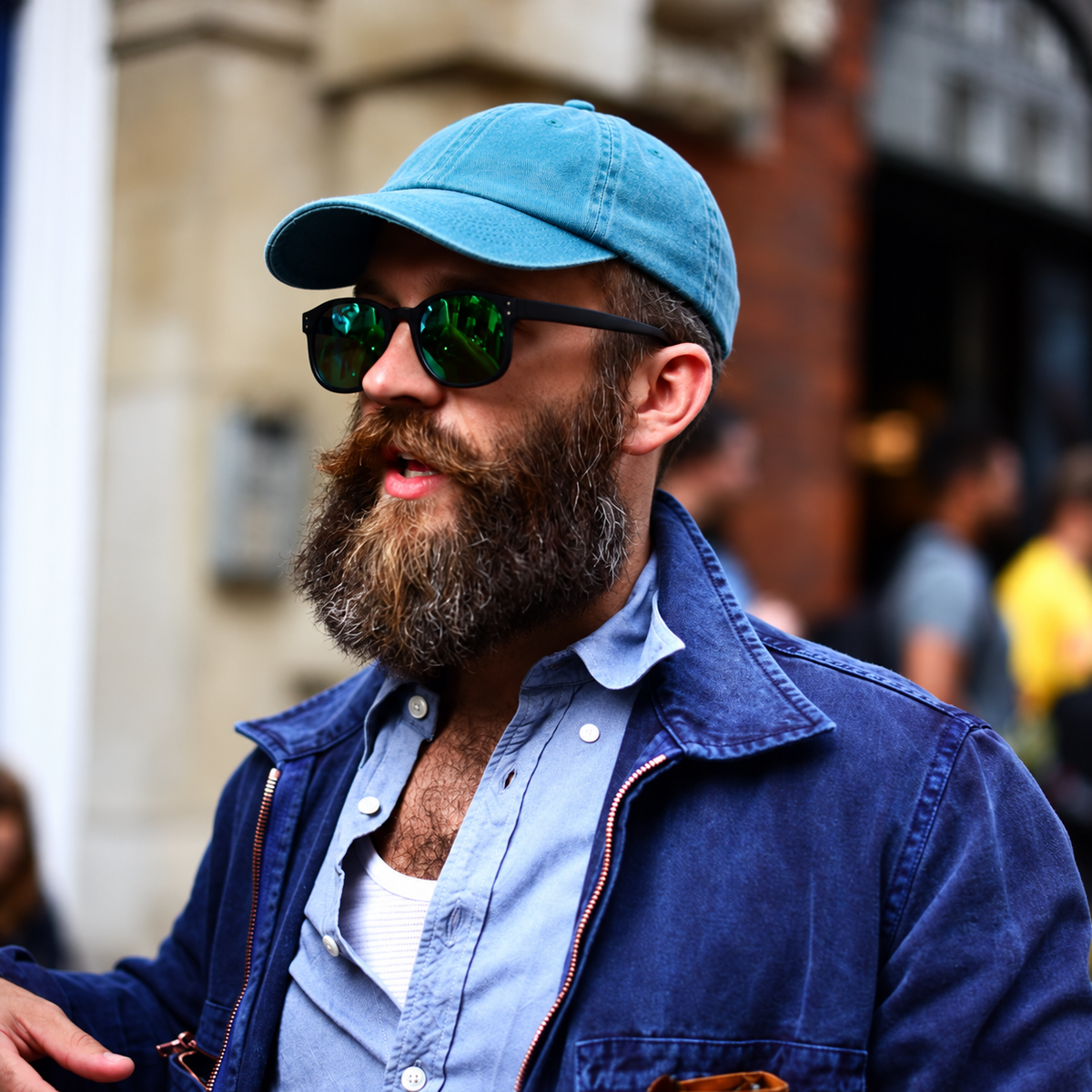 A man with a full beard wearing a light blue baseball cap, black sunglasses with green reflective lenses, a light blue button-up shirt, and a dark blue denim jacket, captured in an outdoor street setting.