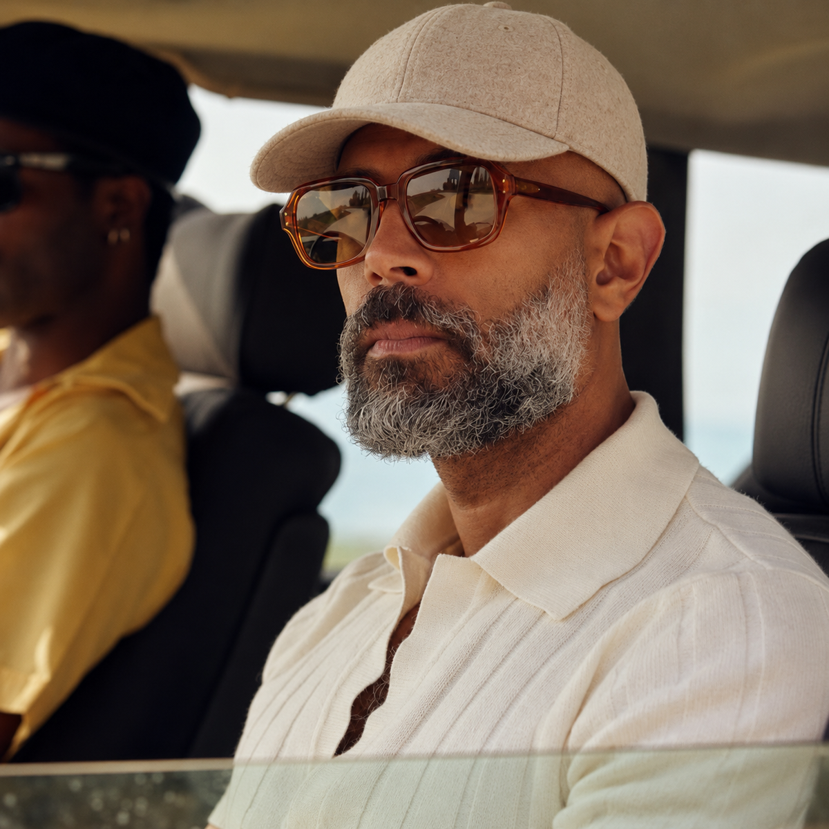 A man wearing a beige baseball cap, brown tortoiseshell sunglasses, and a cream-colored pleated polo shirt, sitting inside a car with another person visible in the background.