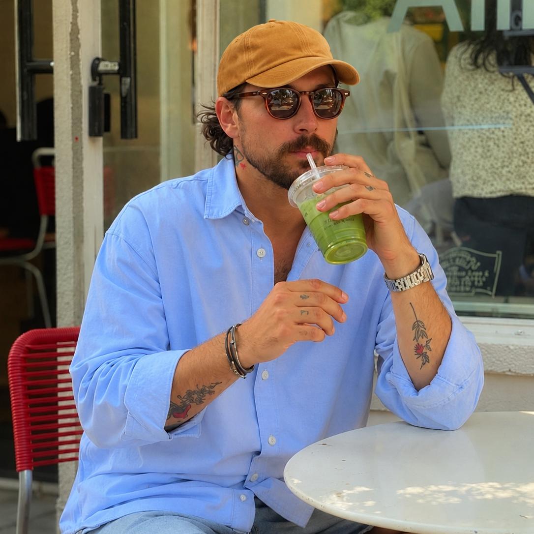 A man wearing a light blue button-down shirt, denim shorts, and a tan baseball cap sits at a white table at an outdoor cafe, drinking a green drink from a clear plastic cup.