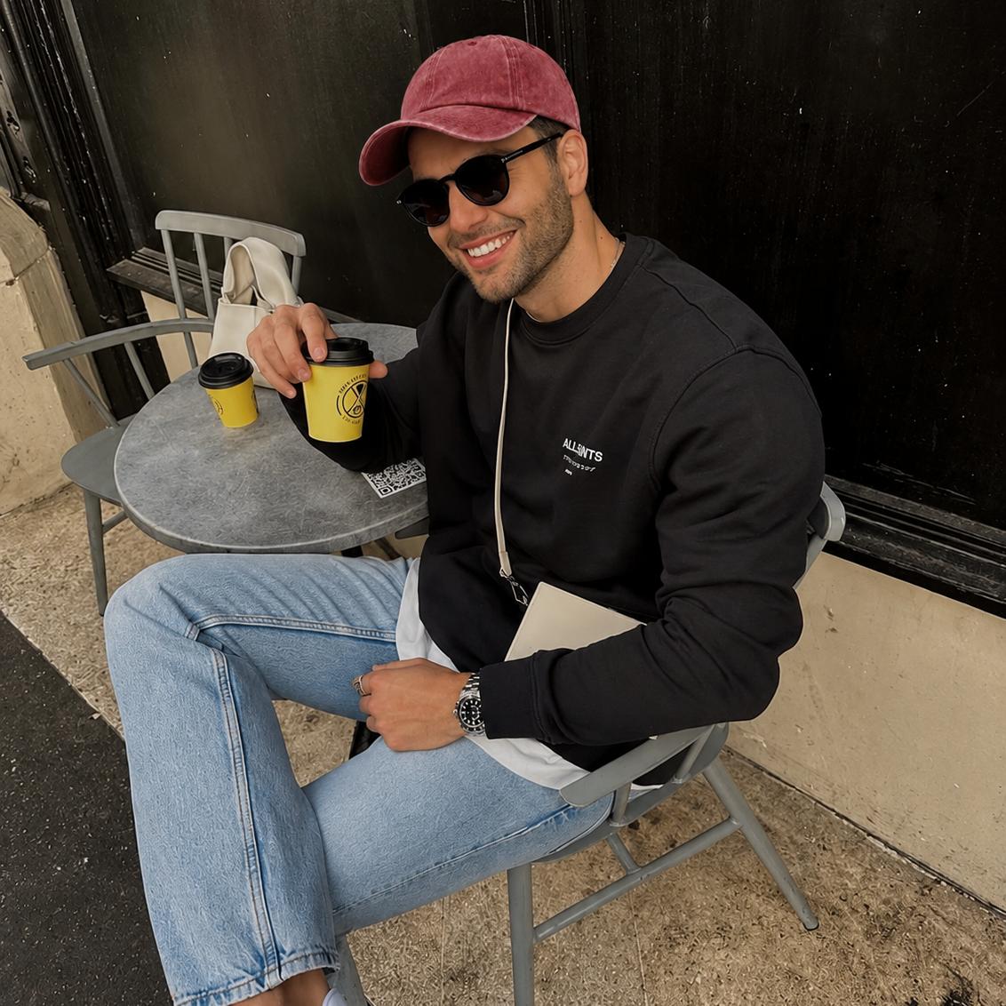 A man wearing a black long-sleeve sweatshirt, light blue jeans, a maroon baseball cap, and sunglasses sits on a metal chair at an outdoor cafe table.