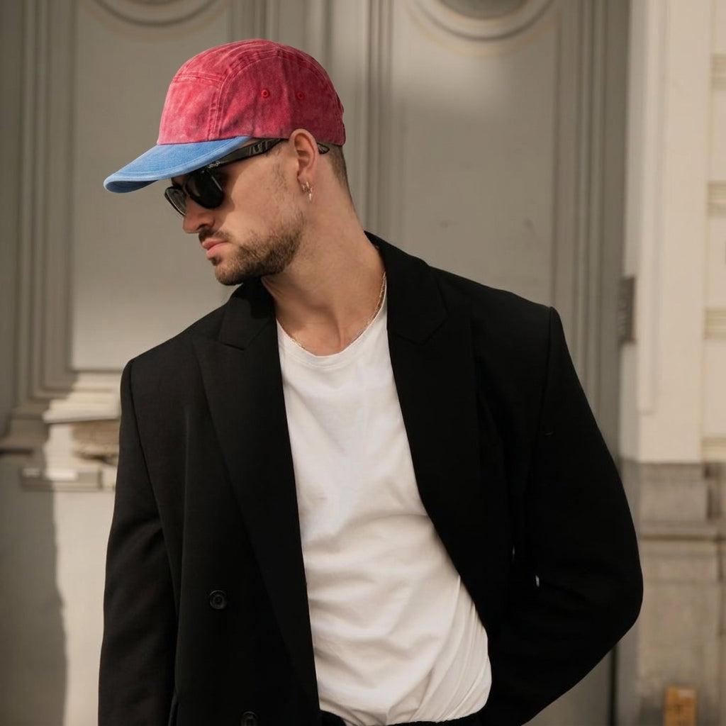 A man wearing a red and blue baseball cap, black sunglasses, a black blazer, and a white t-shirt, photographed in an elegant indoor setting with classical columns.