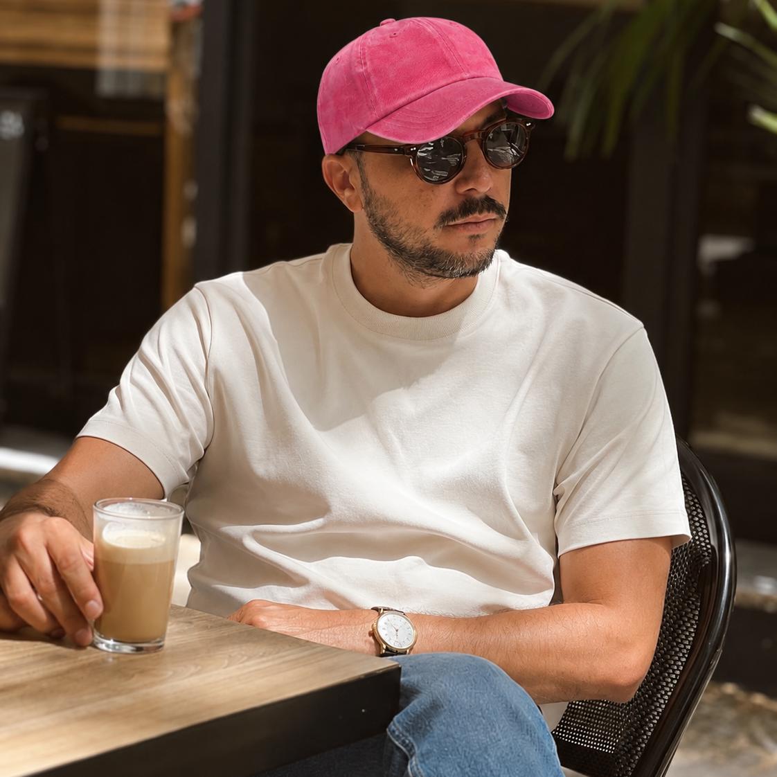 A man sitting at an outdoor cafe table wearing a white crewneck t-shirt, blue jeans, a pink baseball cap, and sunglasses, with a glass of iced coffee on the table.
