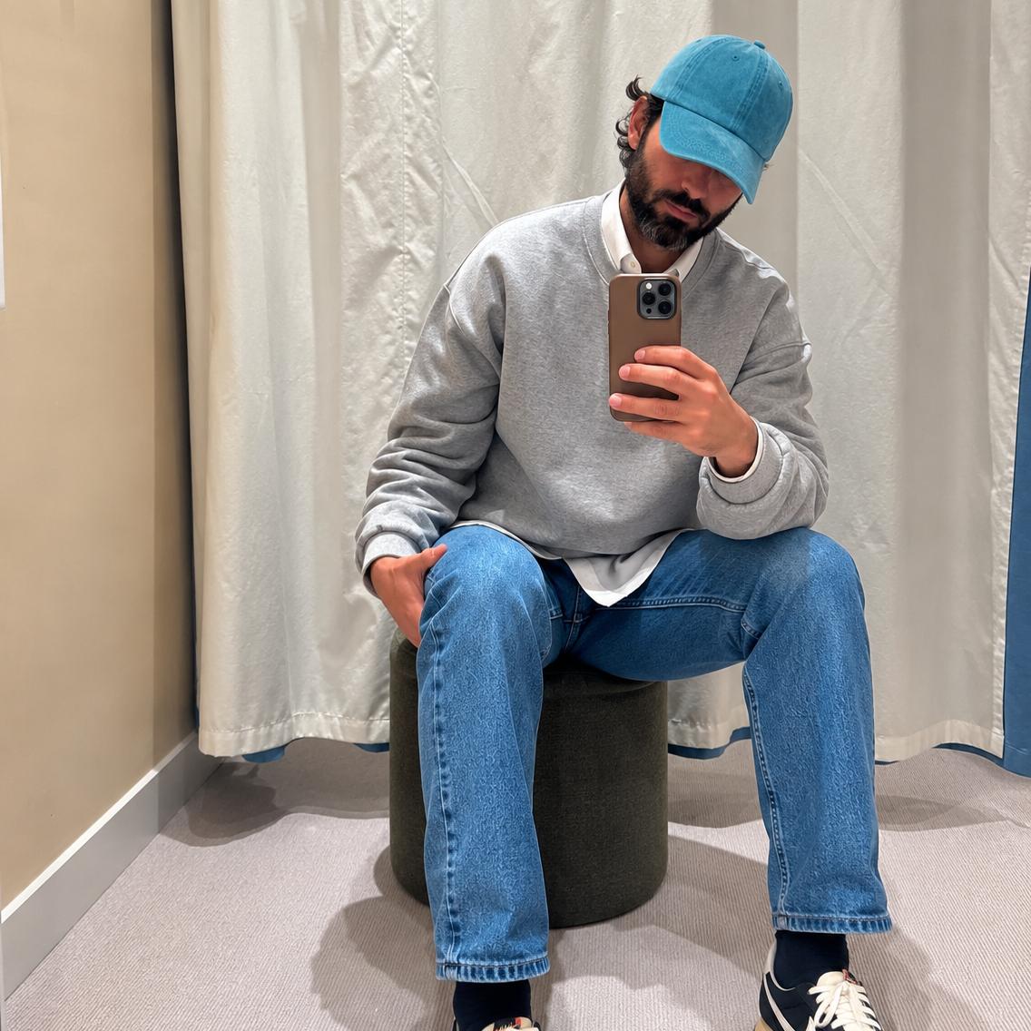 A man takes a mirror selfie in a fitting room wearing a light gray crewneck sweatshirt, blue jeans, a blue baseball cap, and dark blue Nike sneakers with white laces and a white swoosh.