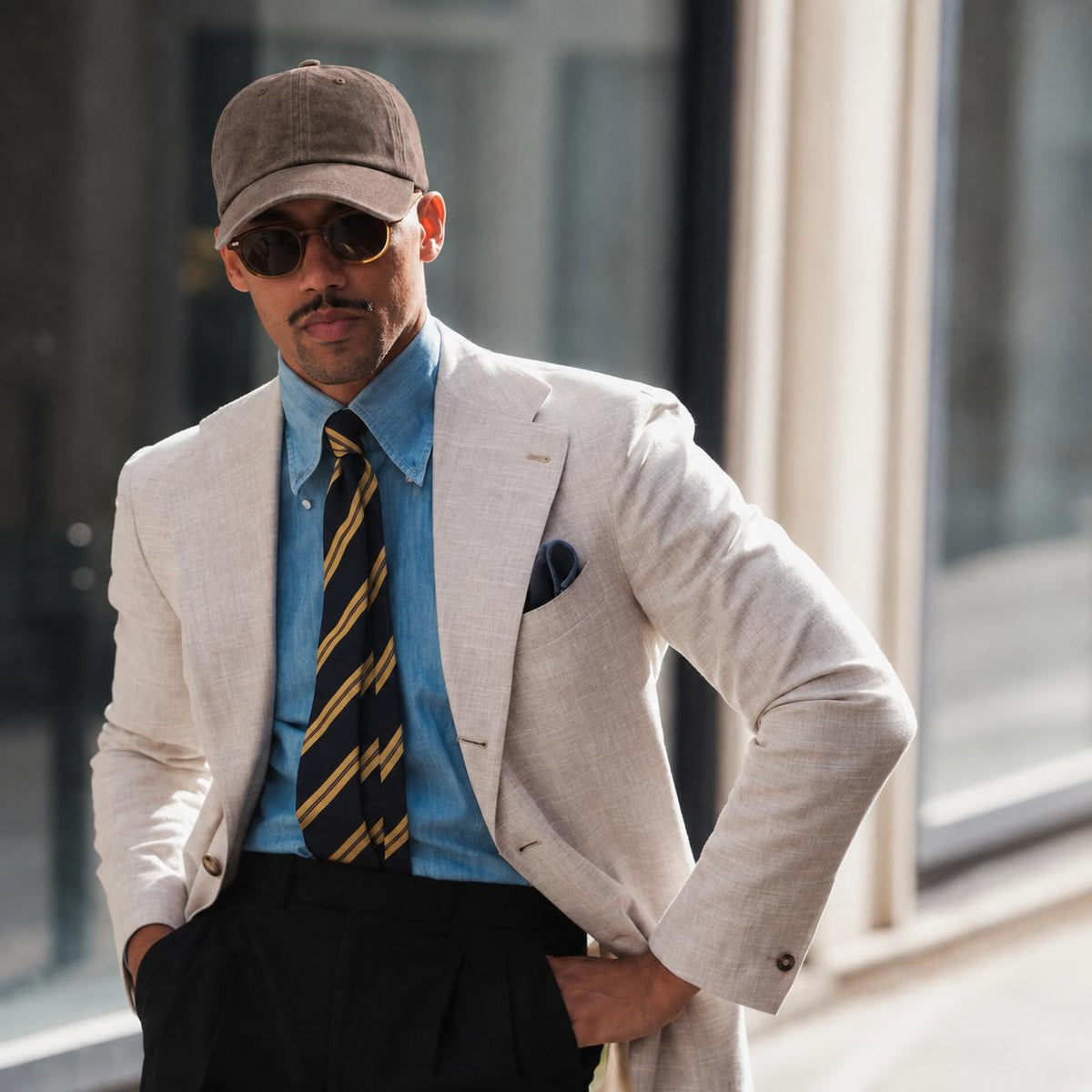 A man wearing a light beige linen blazer, a light blue dress shirt, a yellow and black striped tie, black trousers, a brown baseball cap, and sunglasses, standing in front of a blurred modern building background.