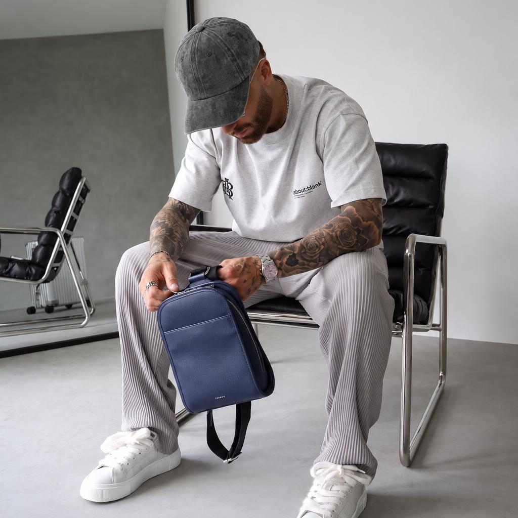 A man wearing a white t-shirt, grey sweatpants, and white sneakers sits on a black chair in a minimalist room, holding a navy blue crossbody backpack.