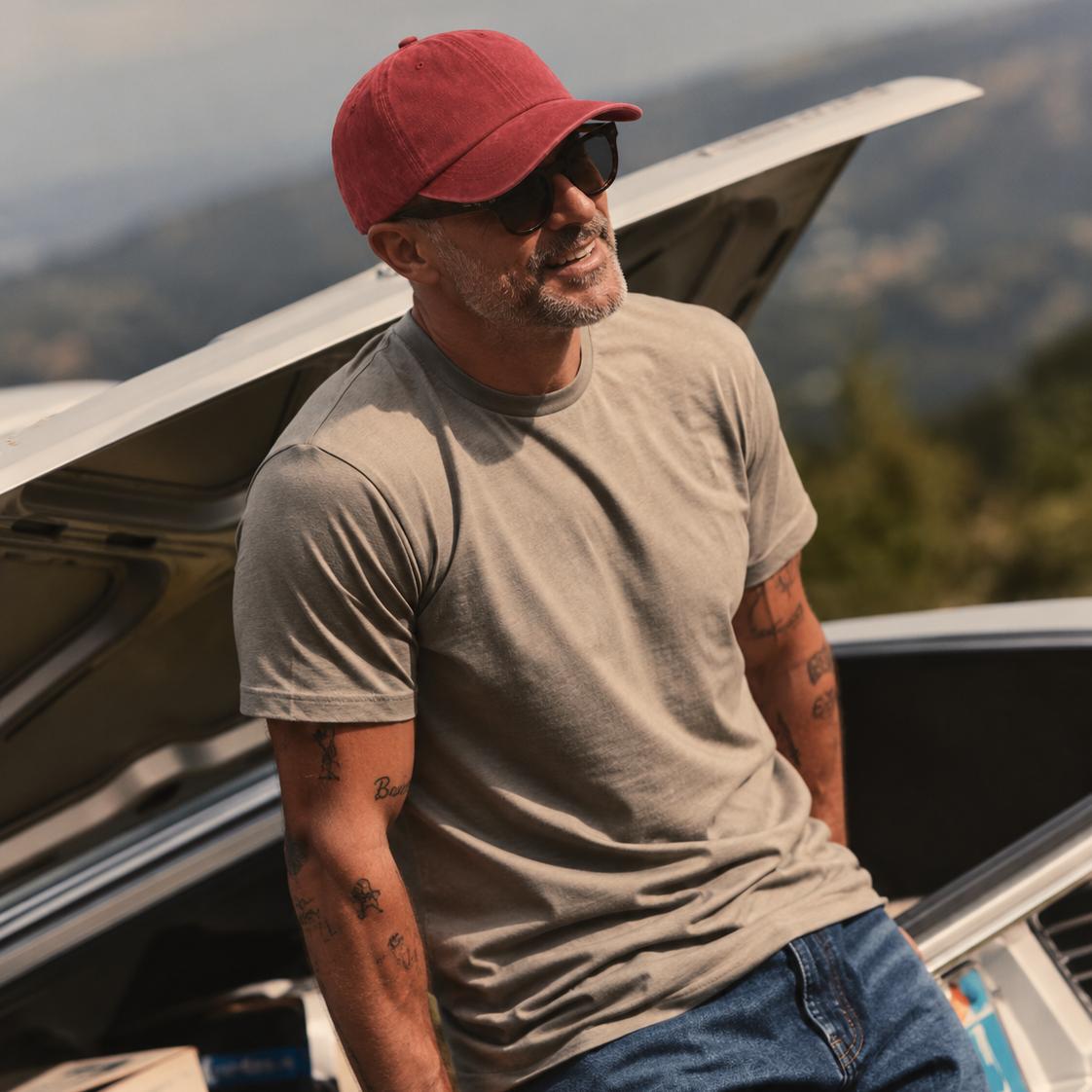 A man wearing a red baseball cap, sunglasses, a beige t-shirt, and blue jeans leans against the open trunk of a white truck outdoors with a blurred mountain landscape in the background.