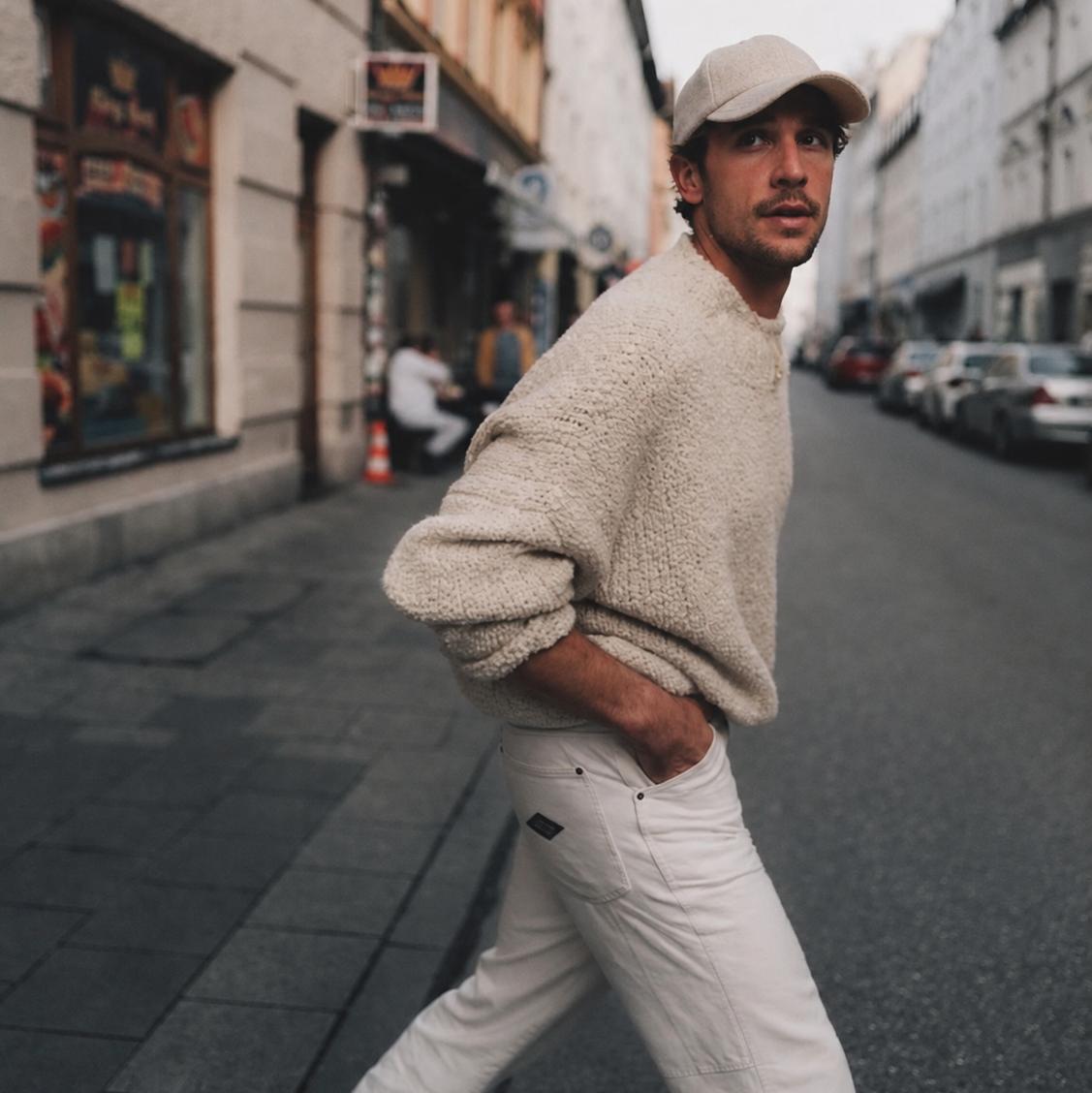 A man walking on a city street wearing a beige sherpa fleece sweater, off-white trousers, a beige baseball cap, and white sneakers.