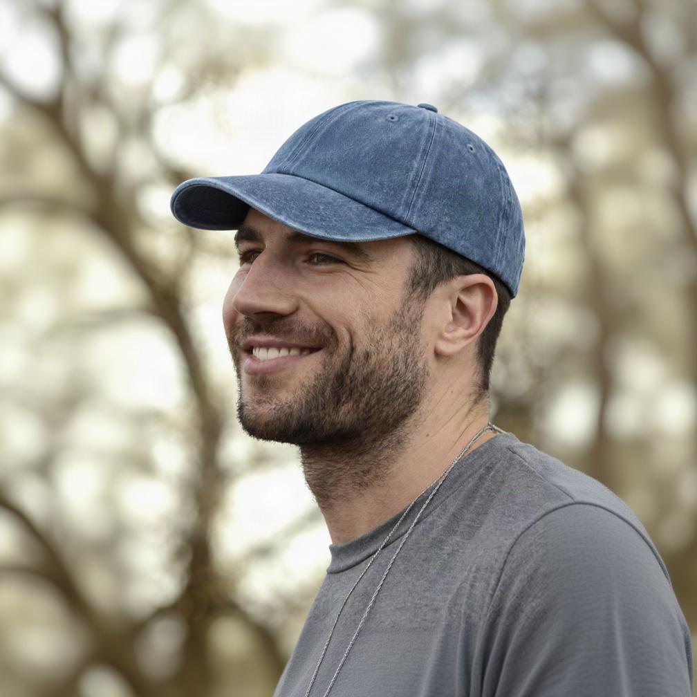 A smiling man in profile wearing a blue denim baseball cap and a gray t-shirt, photographed outdoors with a blurred natural background.