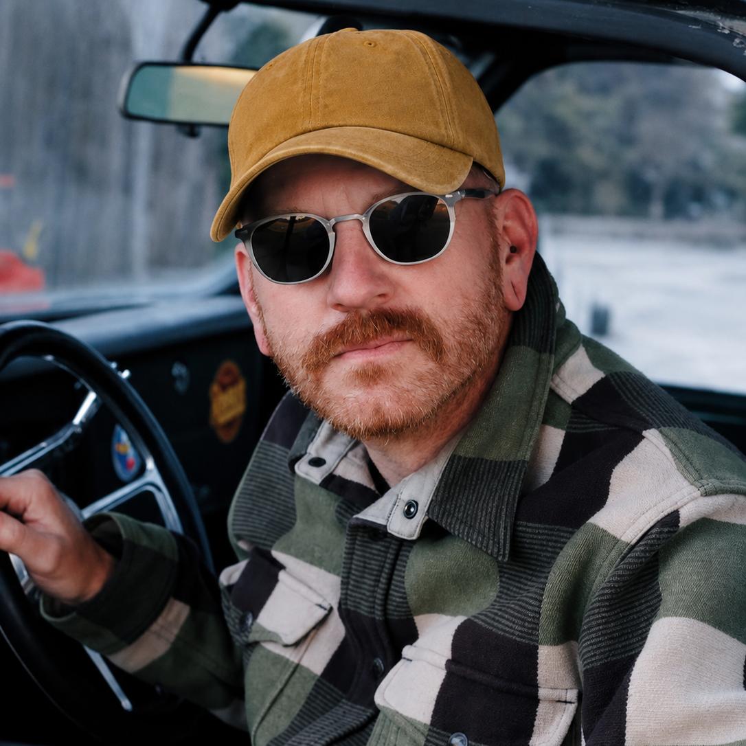 A man wearing a mustard yellow baseball cap, round sunglasses, and a green, black, and beige plaid flannel shirt sits in the driver's seat of a vintage car.