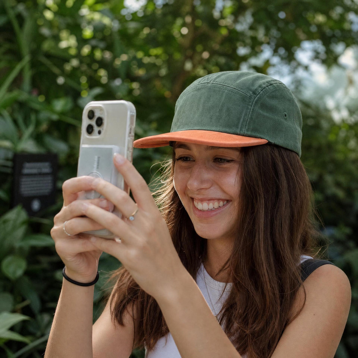 A woman smiling while taking a photo with her phone outdoors, wearing a green and orange baseball cap, a white tank top, and rings.
