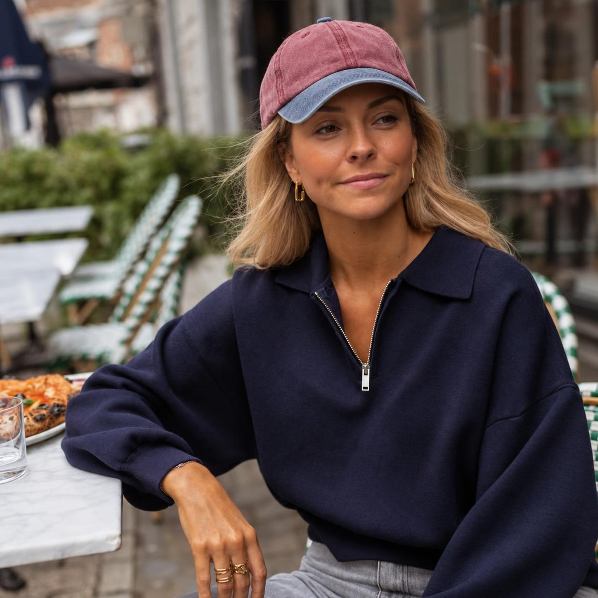 A woman sitting at an outdoor cafe table wearing a navy blue half-zip sweatshirt, gray jeans, and a dusty rose and blue baseball cap.