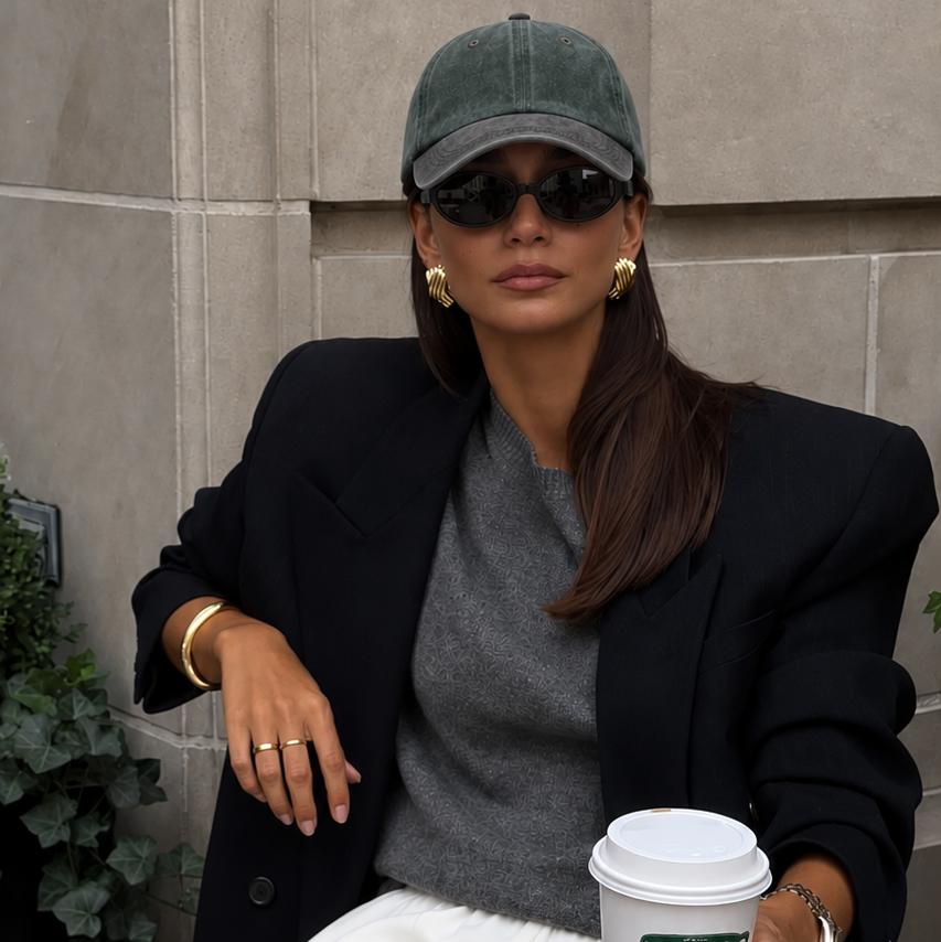 A woman sits at an outdoor cafe table wearing a dark green baseball cap, black sunglasses, a black blazer, a gray top, and white pants, holding a white coffee cup with a green logo.