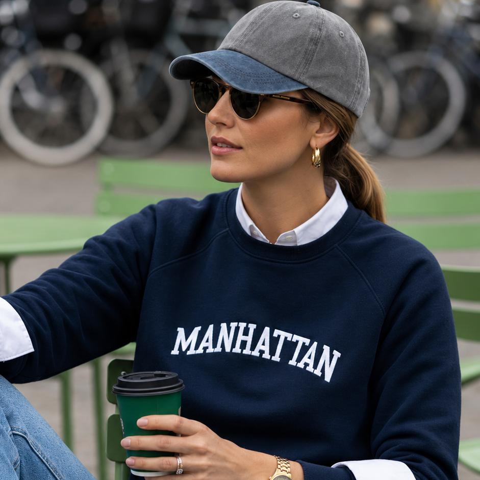 A woman sitting on a green outdoor chair wearing a navy blue crewneck sweatshirt with 'MANHATTAN' printed in white across the chest, layered over a white collared shirt. She is also wearing a gray baseball cap, sunglasses, blue jeans, white socks, and white sneakers, while holding a green coffee cup.