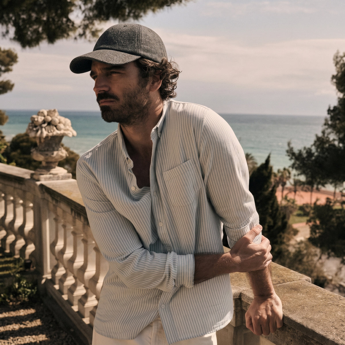 A man wearing a light beige and white vertically striped button-down shirt and a gray baseball cap, leaning on a stone balustrade with the ocean and trees in the background.
