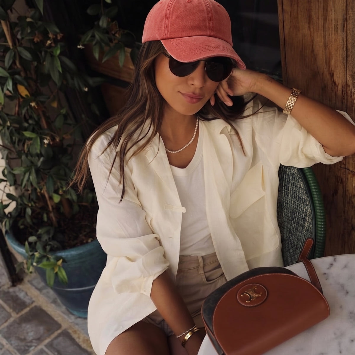 A woman sitting at an outdoor cafe wearing a pink baseball cap, black sunglasses, a cream button-up shirt over a white top, beige shorts, and holding a brown leather crossbody bag.
