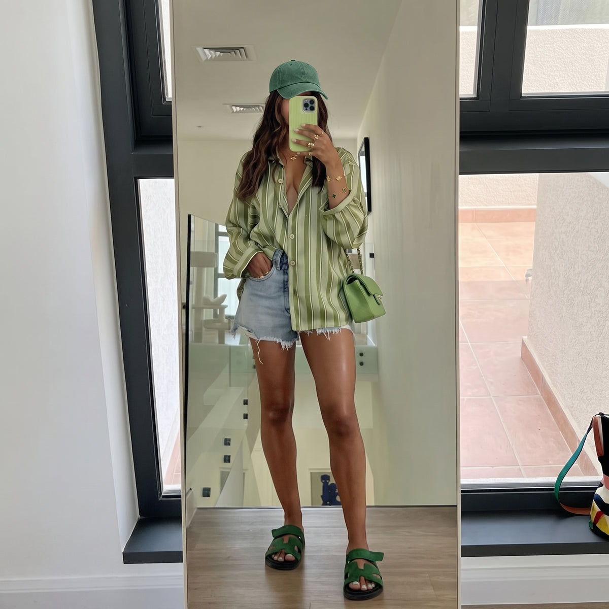A woman takes a mirror selfie in a modern indoor space, wearing a green and white striped button-up shirt, denim shorts, green slide sandals, a green cap, and carrying a small green shoulder bag.