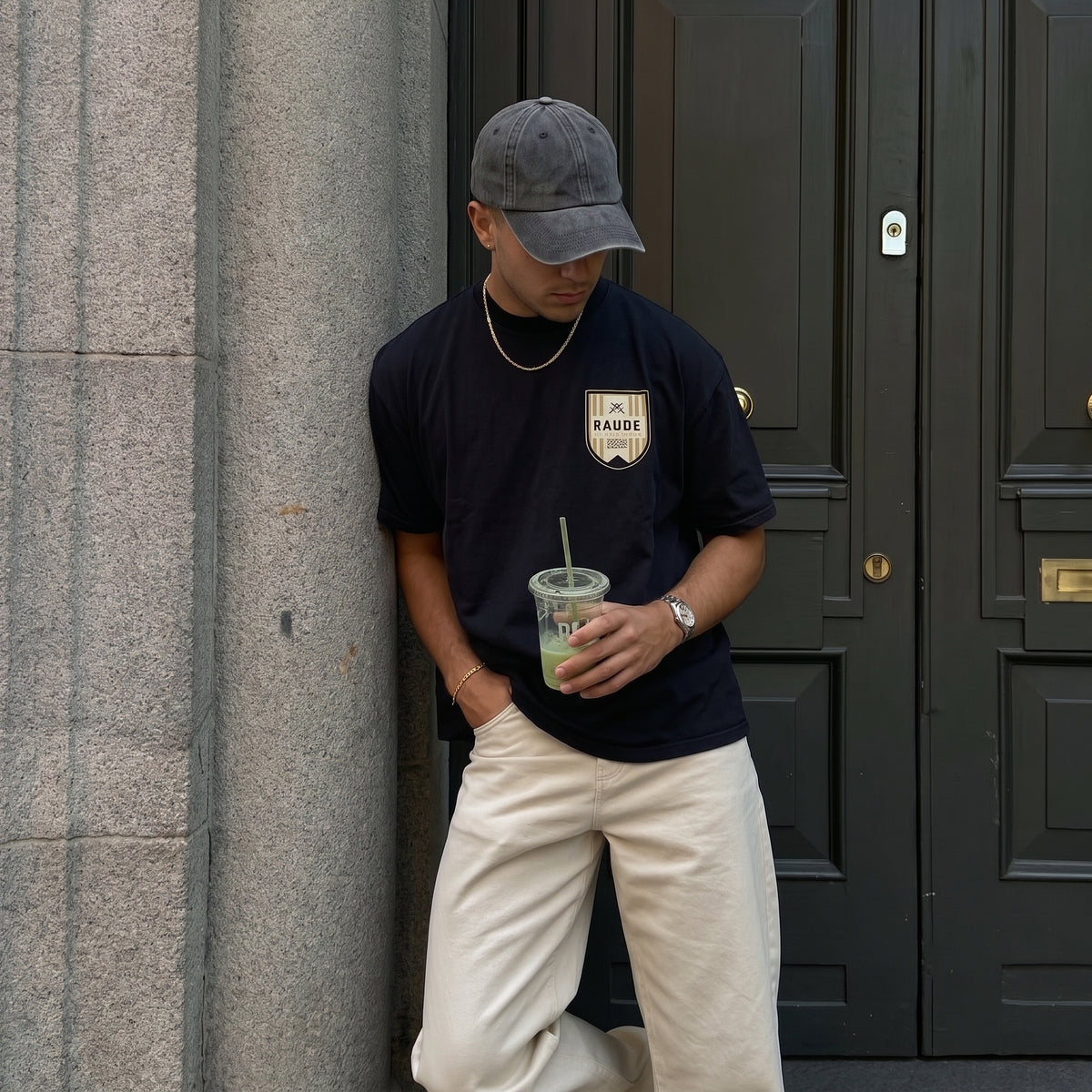 A man wearing a navy blue BADEI t-shirt, beige pants, and a gray baseball cap stands in front of a gray stone column and dark green doors, holding a drink.