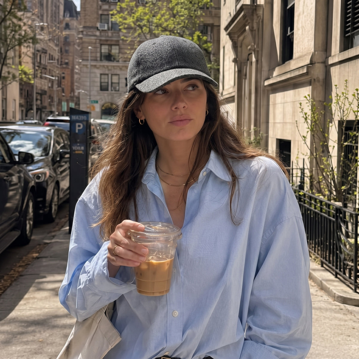 A woman wearing a gray textured baseball cap and an oversized light blue button-down shirt stands on a city street holding an iced coffee.
