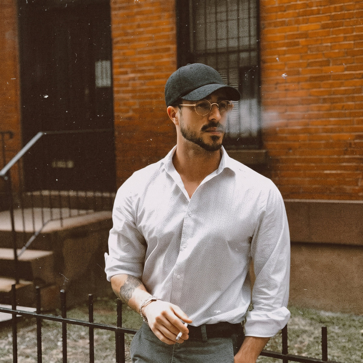 A man wearing a white long-sleeve button-down shirt, dark gray baseball cap, and glasses, standing outdoors in front of a brick building.