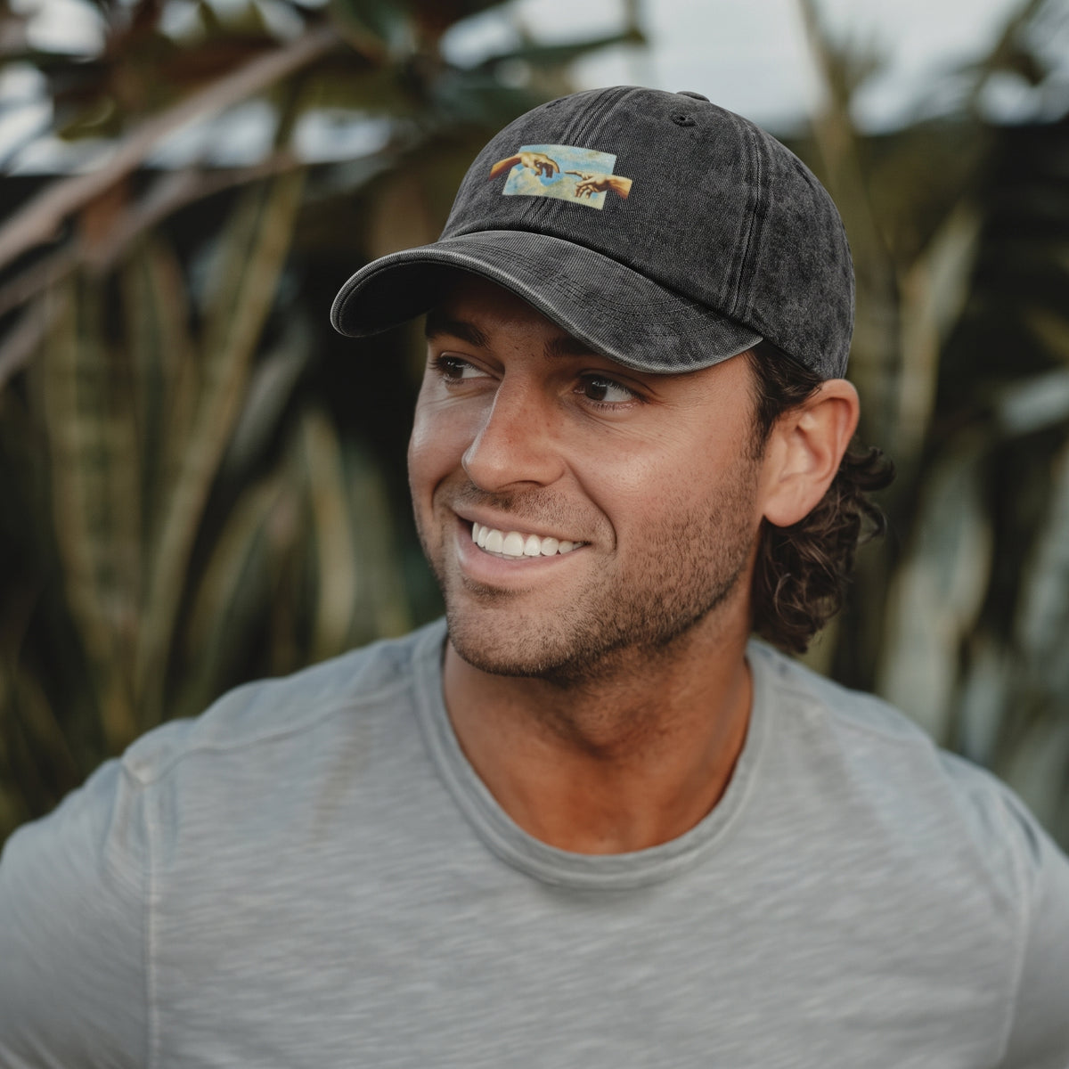 A smiling man wearing a dark gray washed baseball cap with a small colorful graphic patch on the front, photographed outdoors with blurred tropical foliage in the background.
