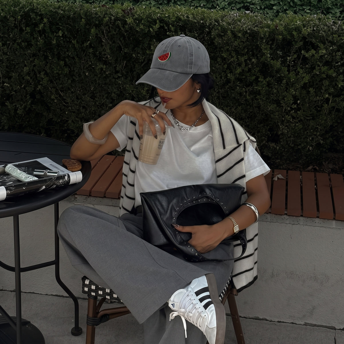A person sitting outdoors at a cafe table wearing a gray baseball cap with a red strawberry patch, a white t-shirt, a black-and-white striped cardigan, gray sweatpants, and white sneakers. They are holding a black studded leather bag and a clear drink.