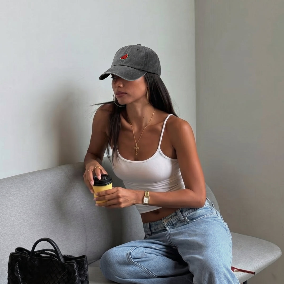 A woman wearing a gray baseball cap with a small red embroidered logo, a white tank top, and light blue jeans sits on a gray sofa holding a yellow travel tumbler. A black handbag is in the foreground.