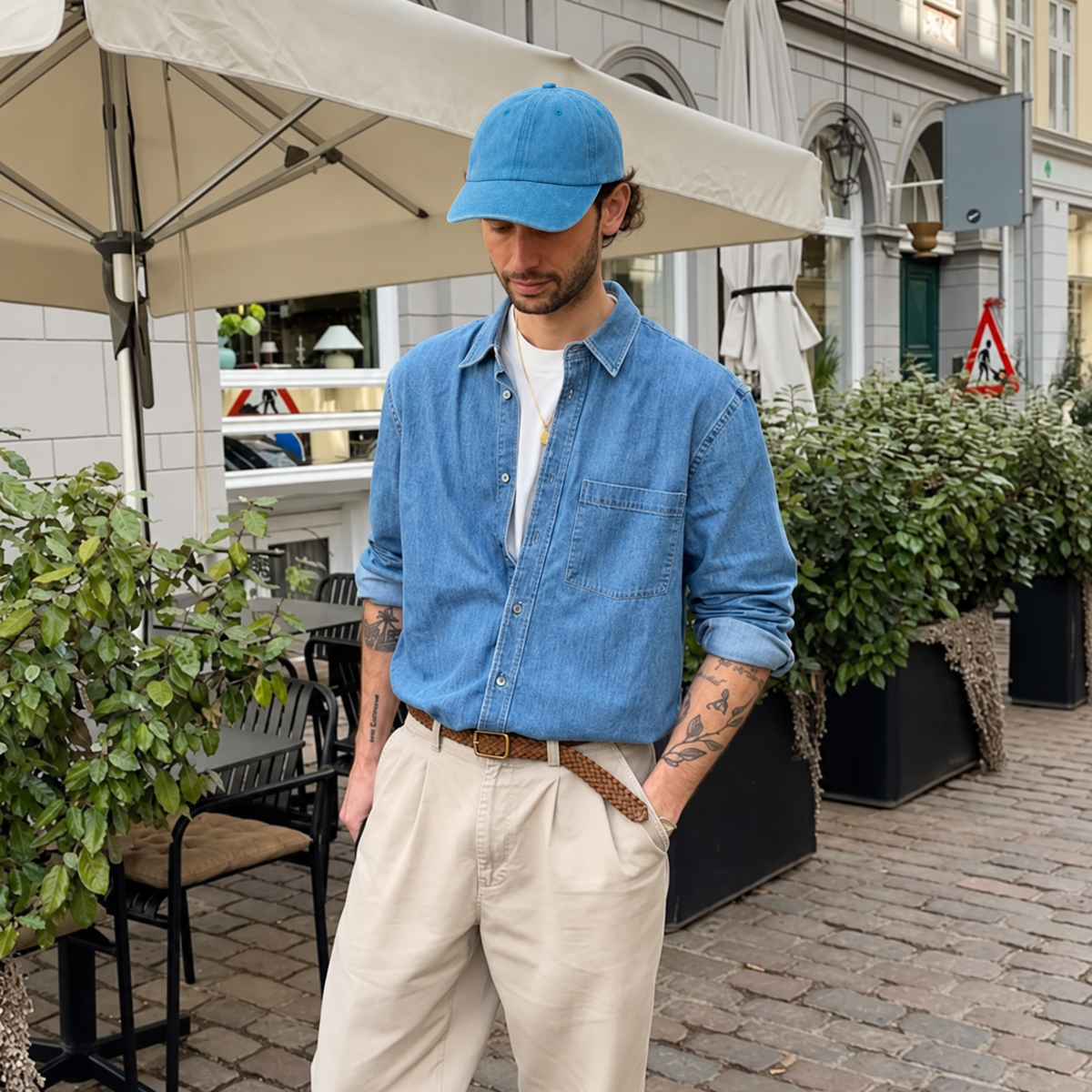 A man wearing a light blue denim chambray shirt, beige trousers, a brown braided belt, and a blue baseball cap, standing on a cobblestone street in front of an outdoor cafe patio.