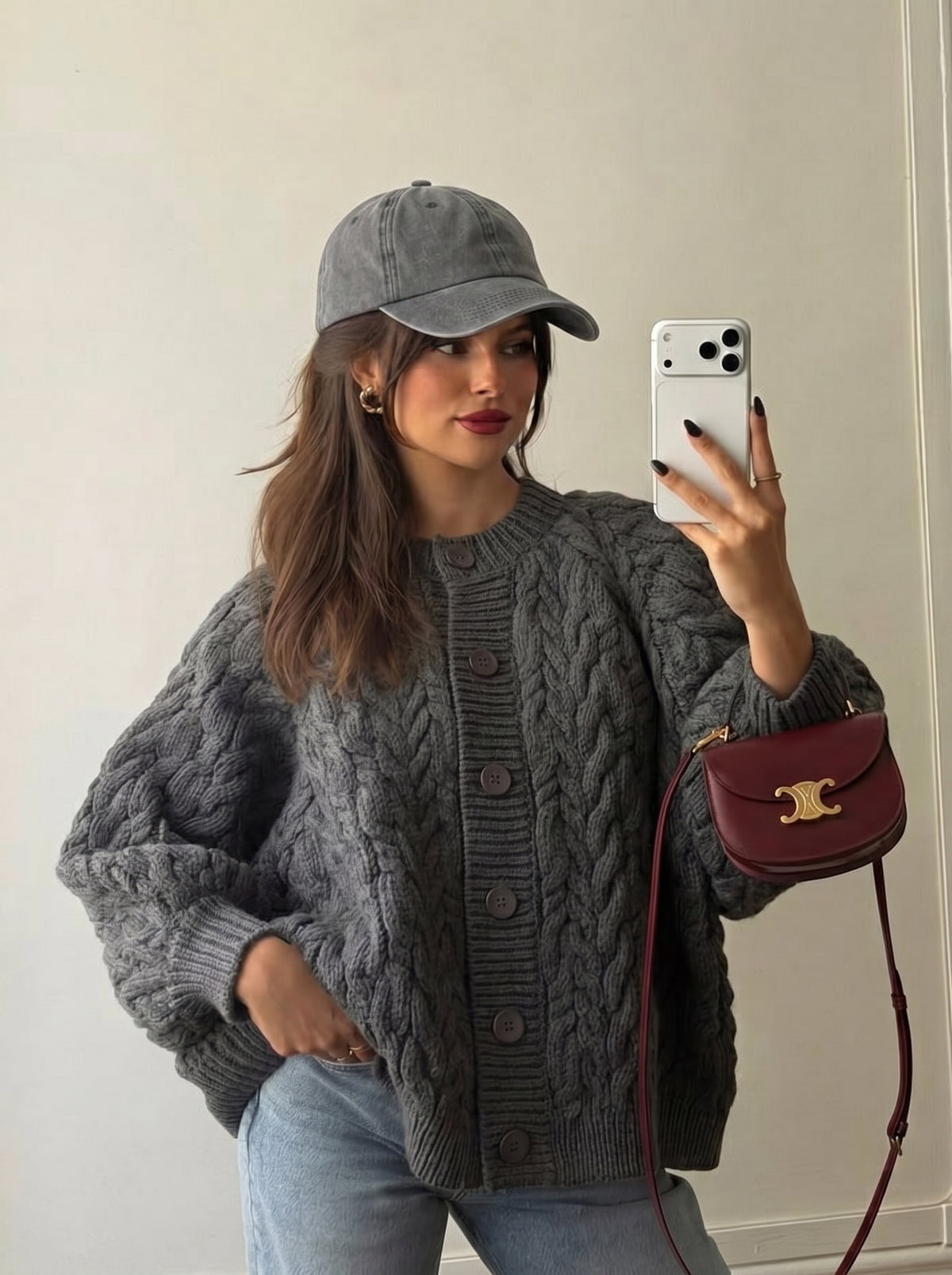 A woman takes a mirror selfie wearing a chunky gray cable-knit button-up cardigan, a gray baseball cap, and light blue jeans, while holding a burgundy crossbody bag with a gold logo clasp.