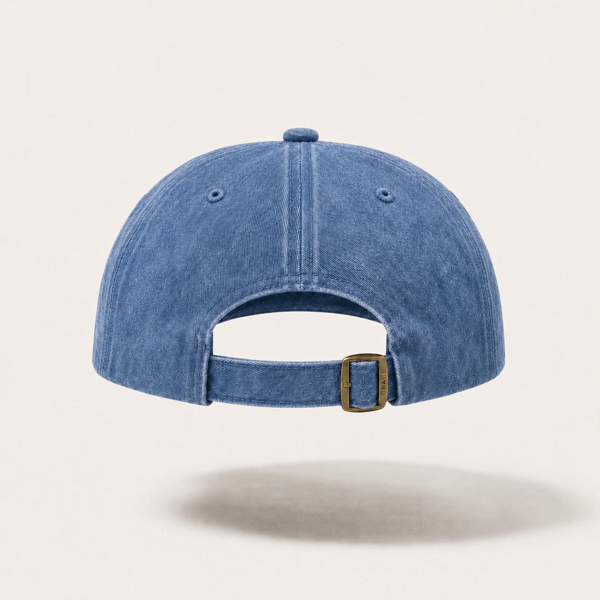 A blue denim baseball cap shown from the back, featuring a curved brim, an adjustable strap, and a small gold-tone metal buckle, set against a light beige background.