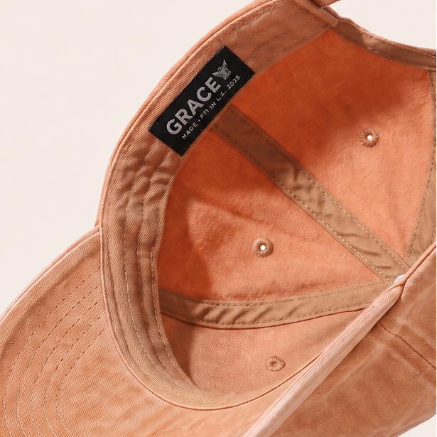 A peach-colored cotton twill baseball cap shown floating at an angle against a warm off-white background, with a black 'GRACE' brand label visible on the inner side.