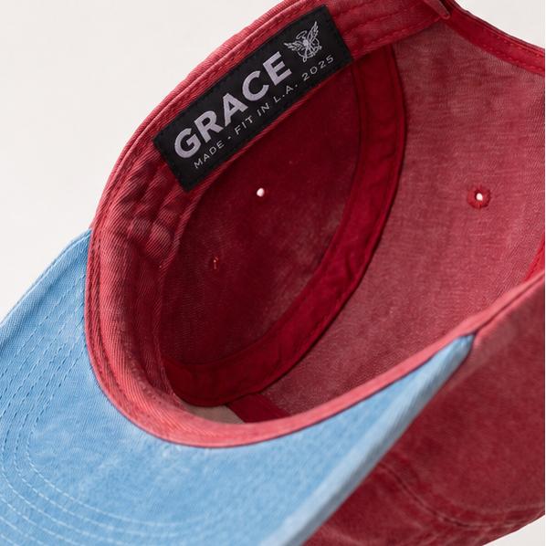 A two-tone baseball cap shown from the side, featuring a washed red crown and a light blue visor. The interior label reads 'GRACE' with a small cross logo.