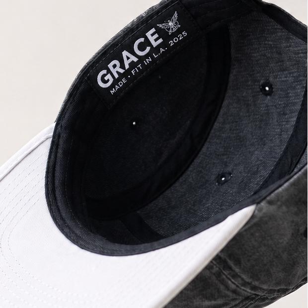A dark washed baseball cap shown floating at an angle, featuring a white curved brim and a black interior with the brand name 'GRACE' printed in white.