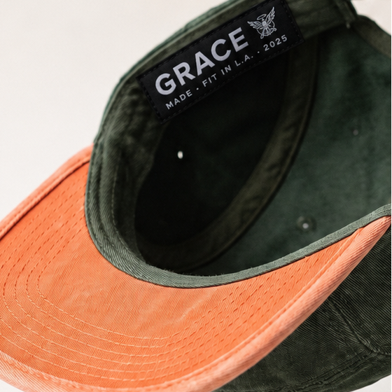 A dark green 5-star baseball cap with a contrasting orange visor, shown floating against a light beige background. The inside of the crown displays the brand name 'GRACE MADE IN LA.