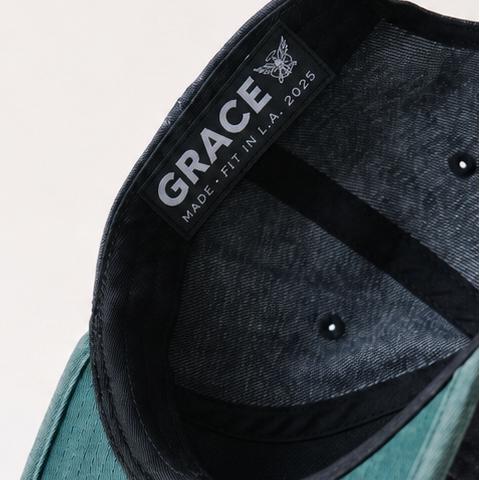 A dark navy blue baseball cap with a teal-green curved brim, shown floating against a light off-white background. The brand name 'GRACE' is visible on the inner side of the crown.