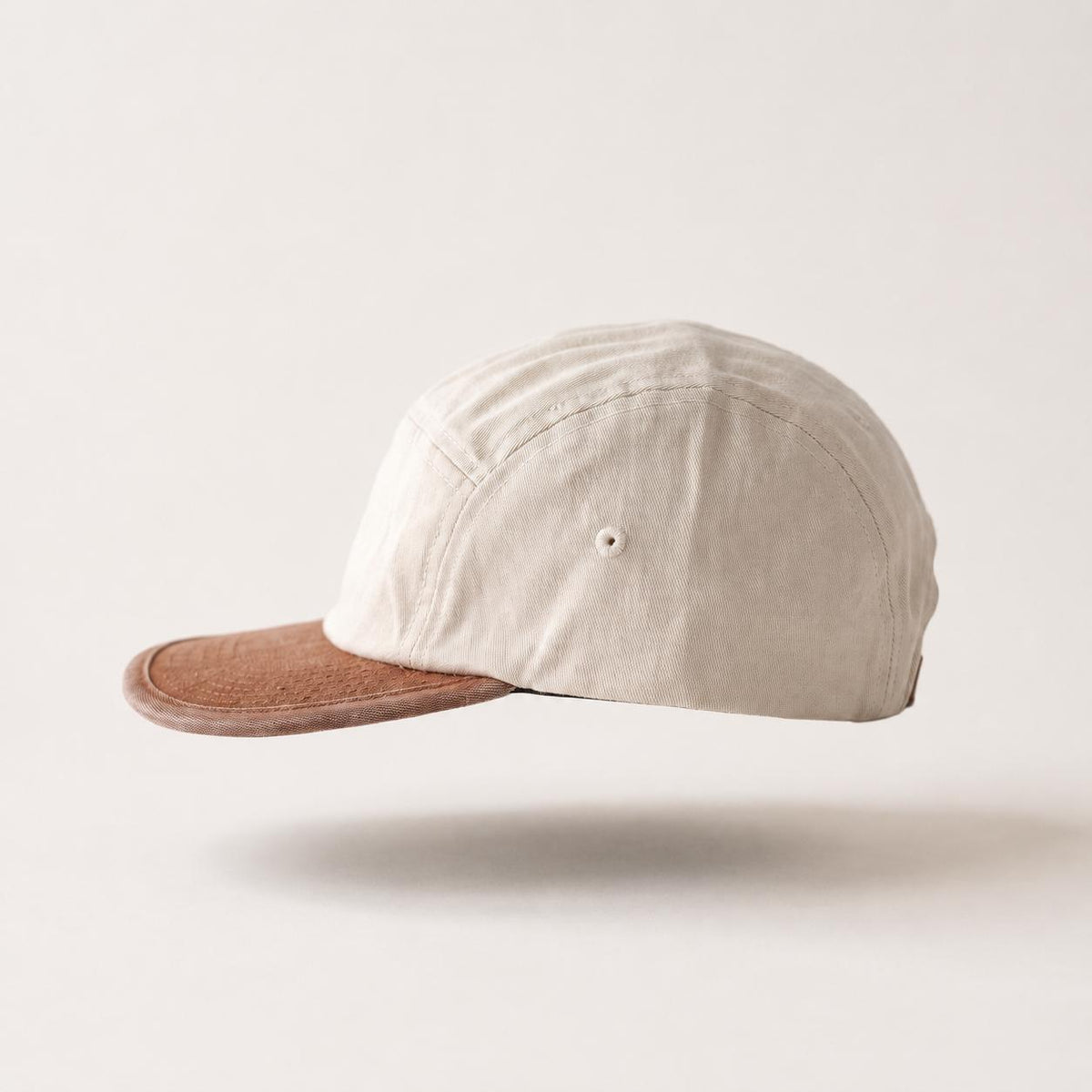 A light beige cotton canvas 5-panel cap with a contrasting brown leather visor, shown floating against a warm off-white background.