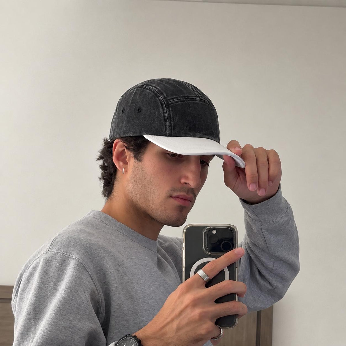 A man taking a mirror selfie wearing a washed black 5-panel baseball cap with a white visor and a gray sweatshirt.