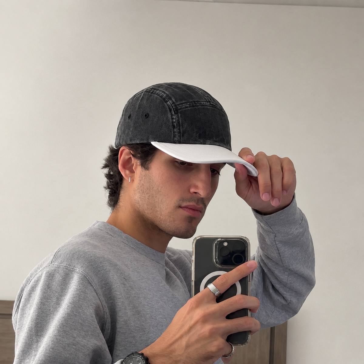A man taking a mirror selfie wearing a washed black 5-panel baseball cap with a white visor and a gray sweatshirt.