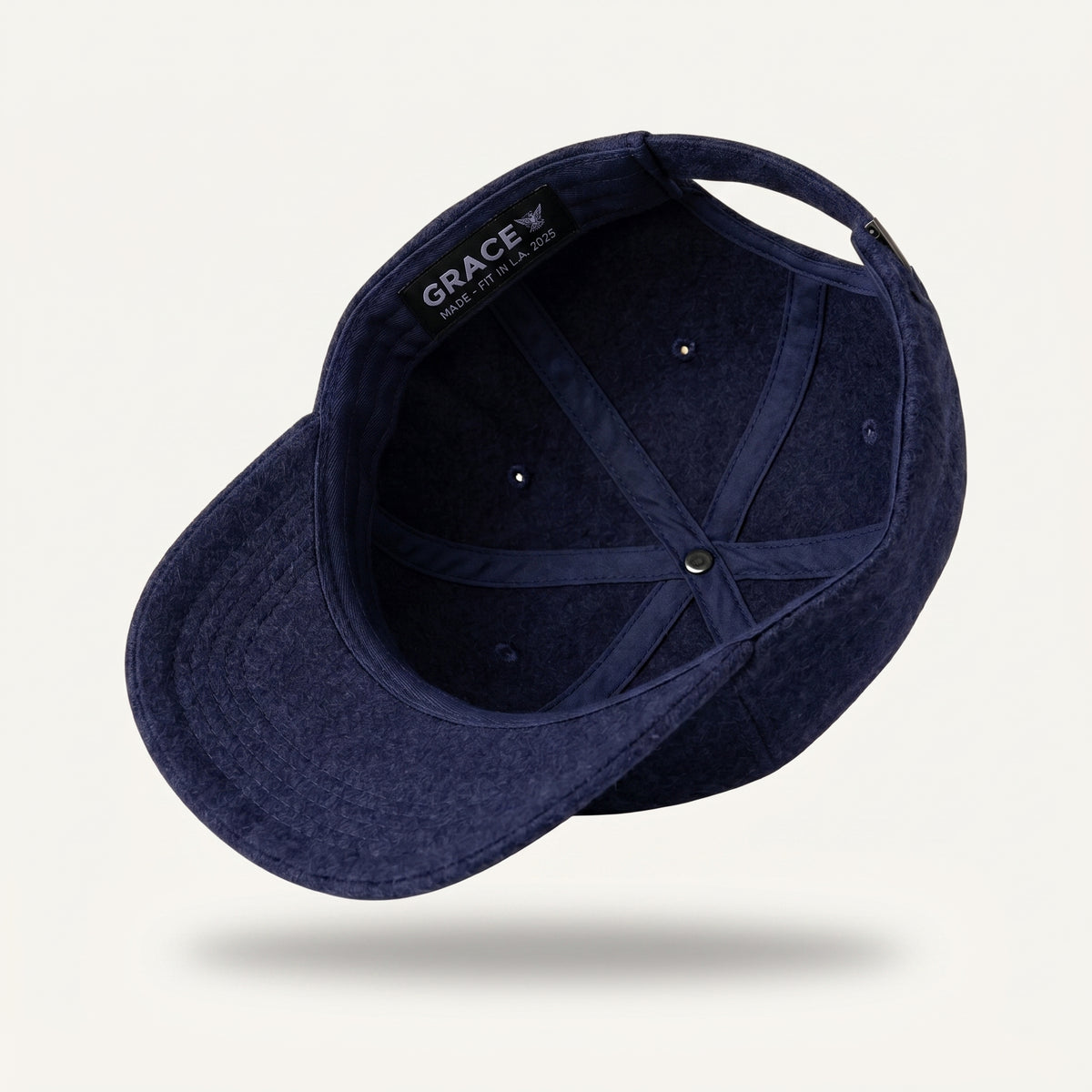 A navy blue textured baseball cap shown from an angled top-down view, floating against a light off-white background with a soft shadow underneath. The brand name 'GRACE' is visible on the inner black label.