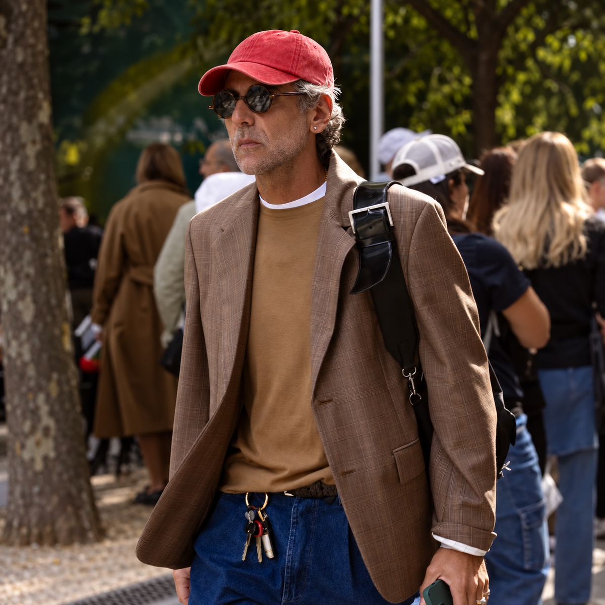 A man walking outdoors wearing a brown blazer, tan sweater, blue jeans, a red baseball cap, sunglasses, and carrying a black shoulder bag.