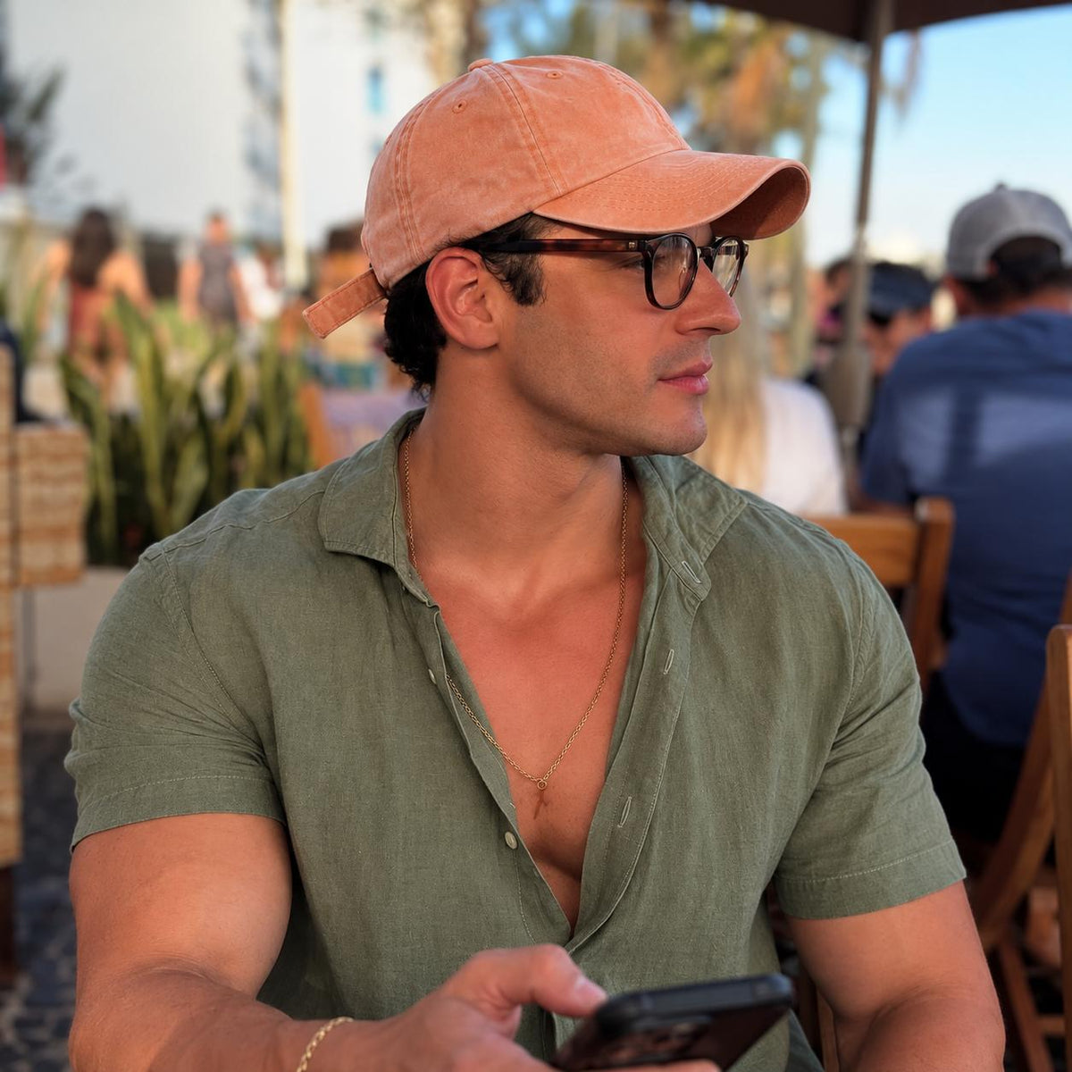 A man wearing a peach-colored baseball cap, black eyeglasses, and an olive green short-sleeve button-up shirt, sitting at an outdoor cafe and looking at his phone.